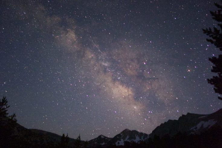 A starry night sky, with the milky way clearly visible in the middle, glowing pink. Around the lower edges, you can see the black outlines of mountains and pine trees.