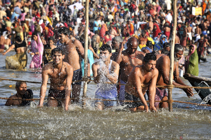 A crowd of people wades in a shallow river. In the background, there is a crowd of hundreds dressed in colorful clothing. In the foreground, a small group of men splashes in the river. On the far left, a man submerges himself completely. The second man from the lefthand side is grinning widely.