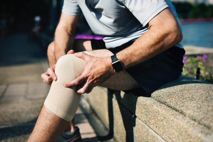 A man, whose head is not visible, sits on a wall and massages his knee, which is wrapped in a brace.