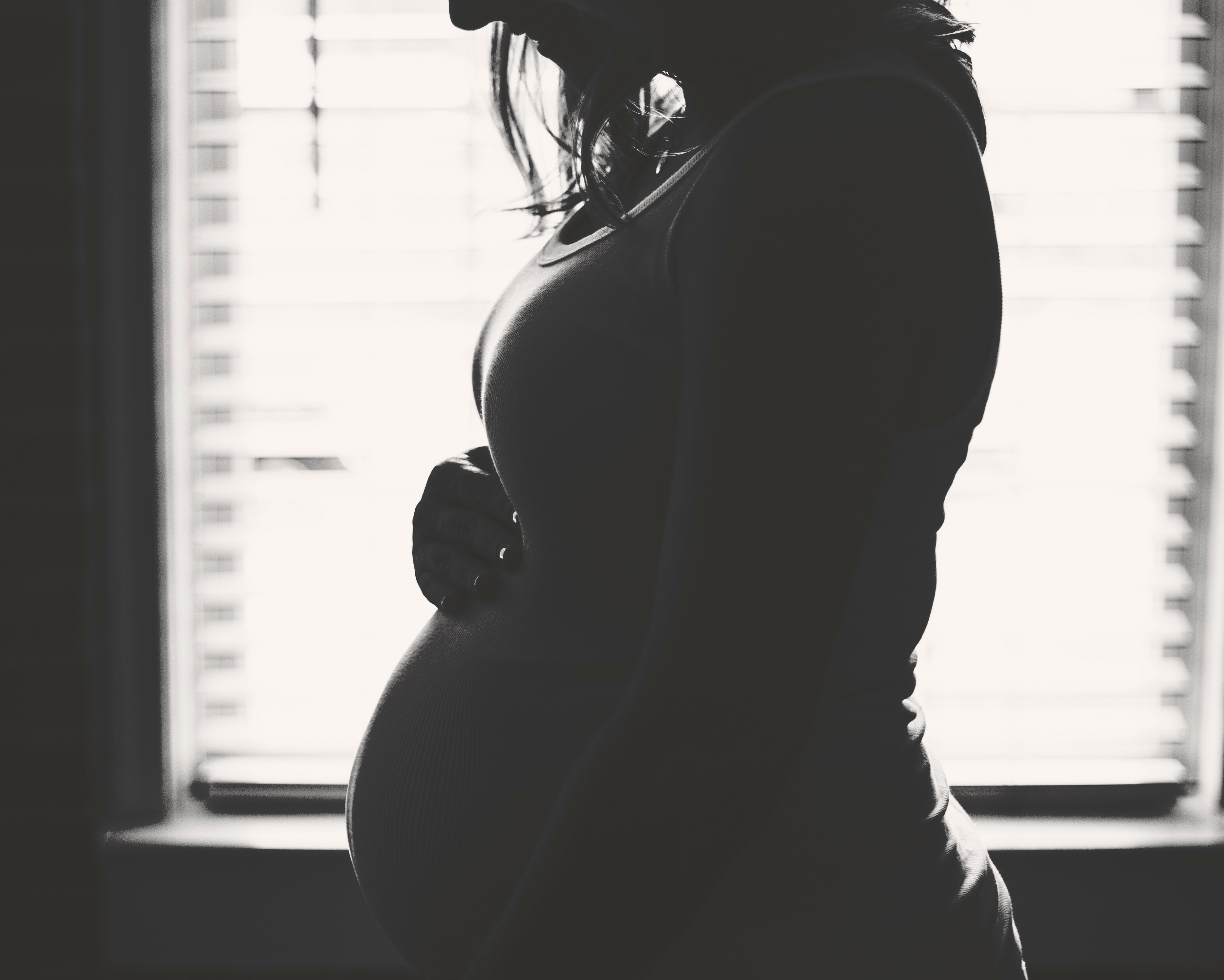 A black and white image of a pregnant woman standing against a window in profile.