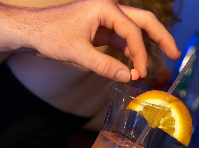 A hand holds a pill above a full glass while a person's back is turned.