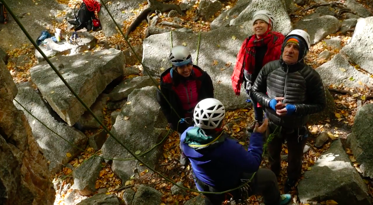 Three climbers look up at an outdoor rockbclimbing route while their instructor explains.