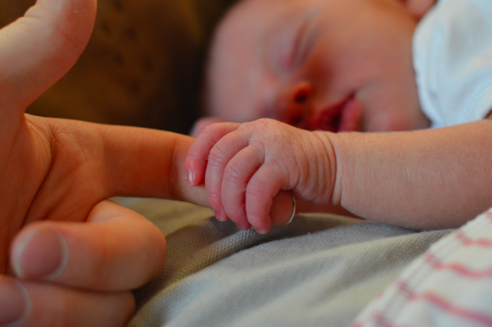 A newborn baby, in the image background, grasps an adults pointer finger, pictured in the foreground