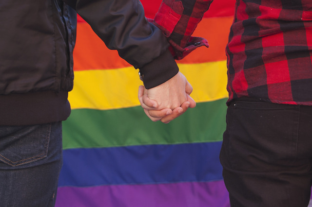 Two men hold hands in front of rainbow flag.