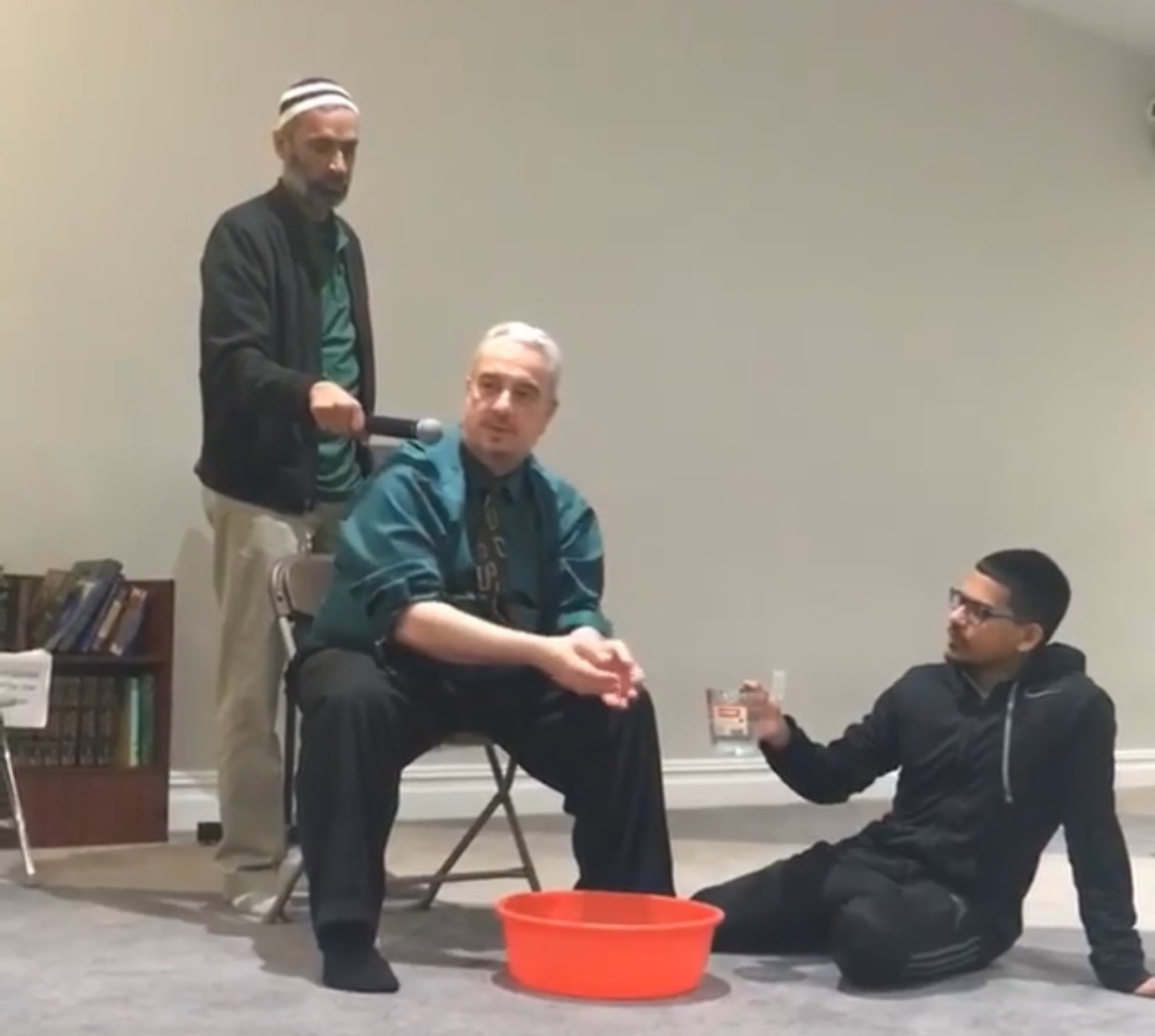 An imam sits in a chair preparing to wash his hands before prayer, while another man stands nearby with a microphone, and a third man sits on the floor holding a cup of water.