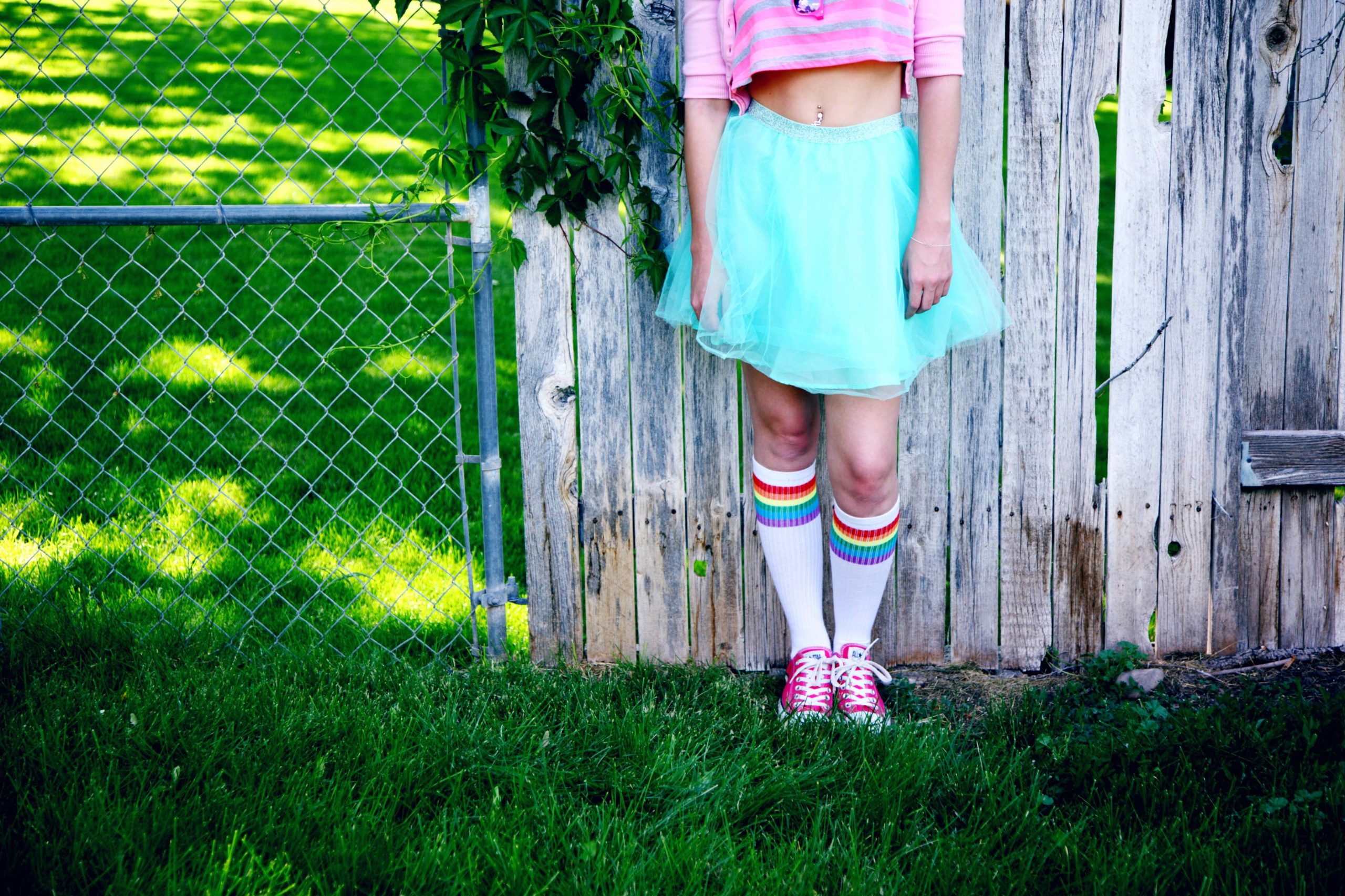 A feminine person stands in front of a wooden fence with a blue skirt, pink crop top and rainbow tube socks