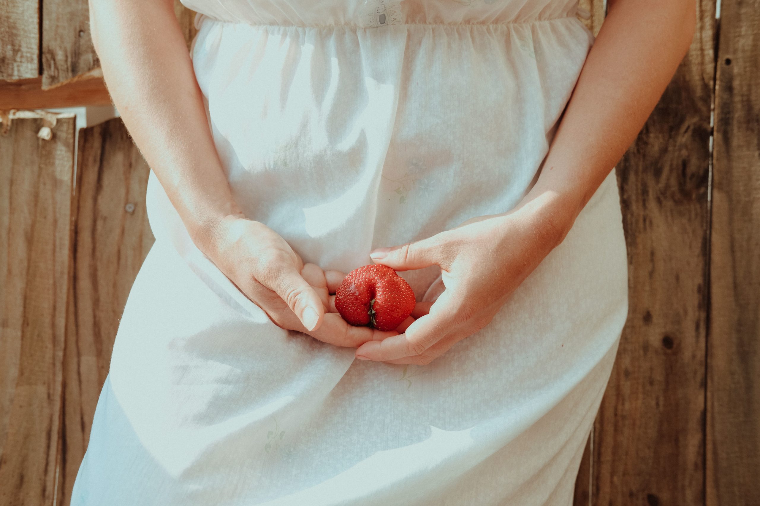 A standing woman in a white dress holds a strawberry in front of her groin.