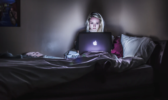 Image of a young women sitting on a bed in dark room on a laptop computer
