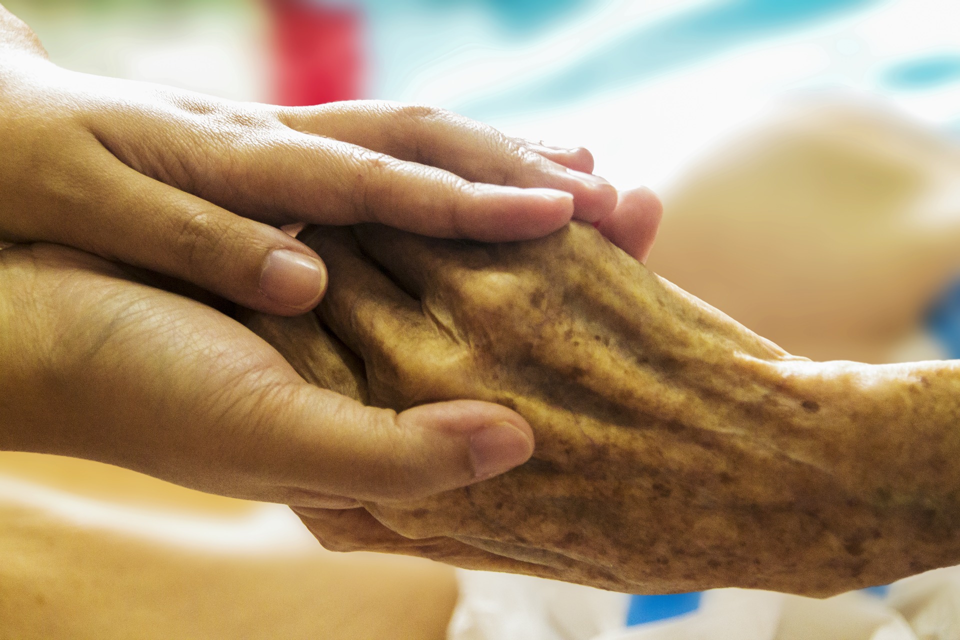 Two smaller hands grasp an older person's hand against the blurred background of a hospital room.