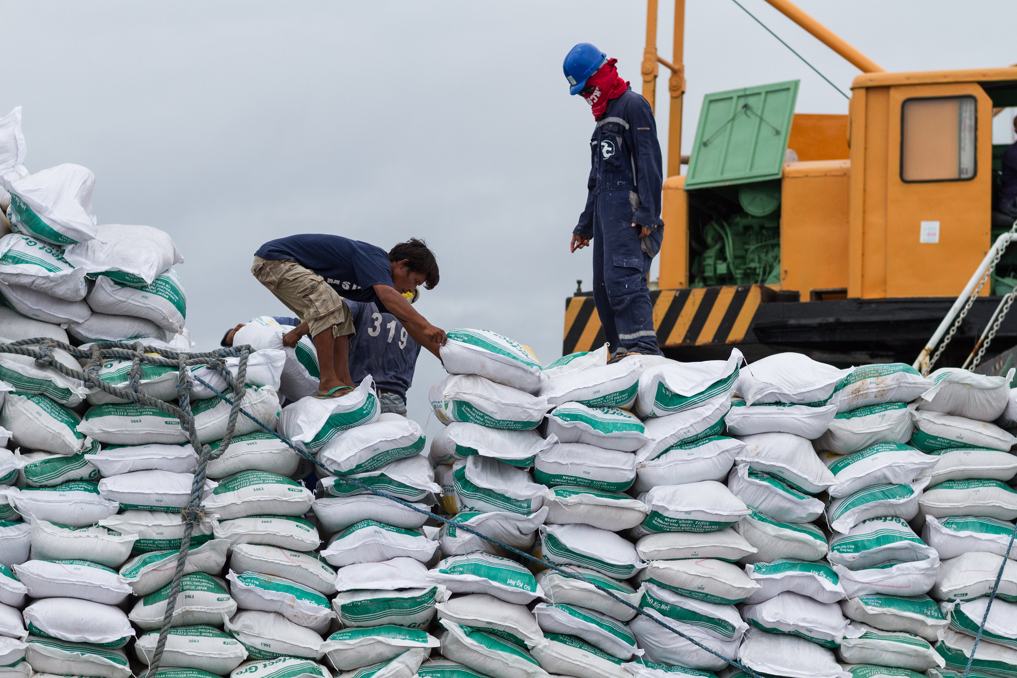 Three workers on top of a large pile of fetilizer bags, with a yellow construction machine behind them.