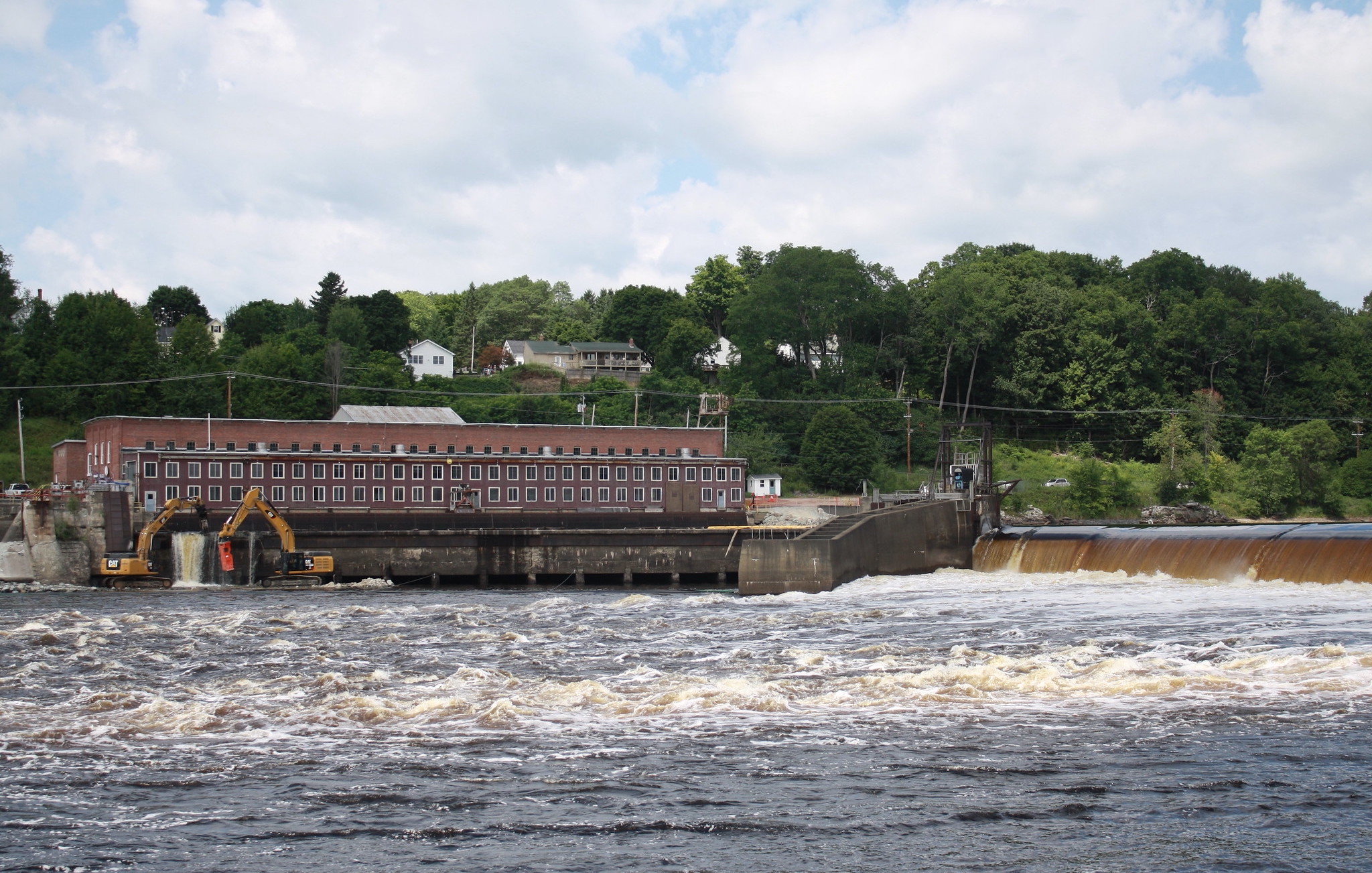 A large brick building sits on top of a big dam, with the river in the foreground. Excavators are demolishing a corner of the dam.