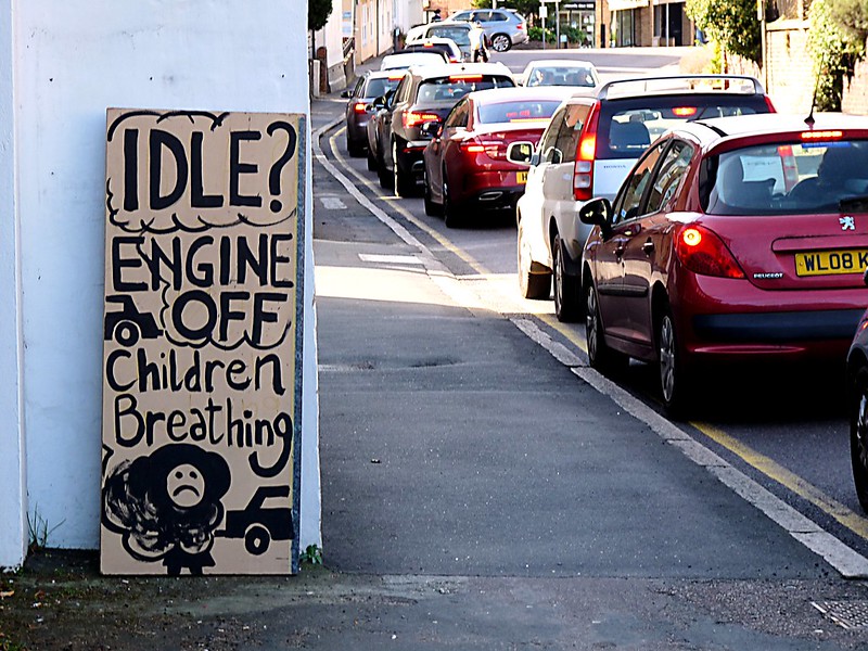 Cardboard sign propped up against a building that reads "Idle? Engine off. Children Breathing." With a drawing of a flower with a frown. Sign is propped up near a line of traffic.