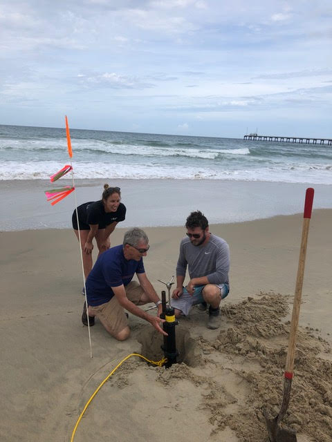 Three scientist at work on a beach deploying a remote sensor. They've dug a three foot deep hole in the sand and are placing a sensor in it.