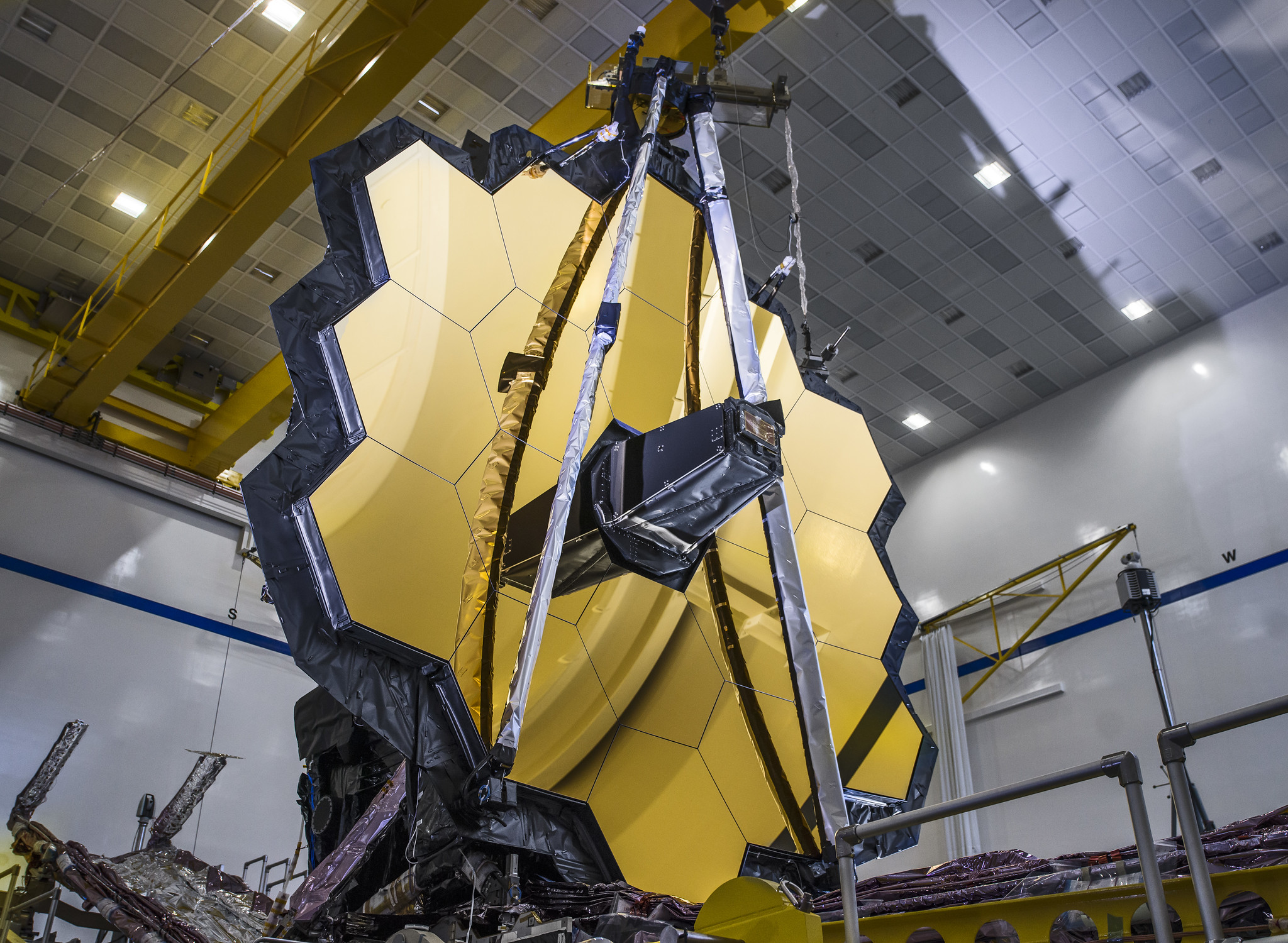 Looking up at the James Webb Space Telescope, assembled inside of a building. It looks like a hexagon of golden, honeycomb mirrors.