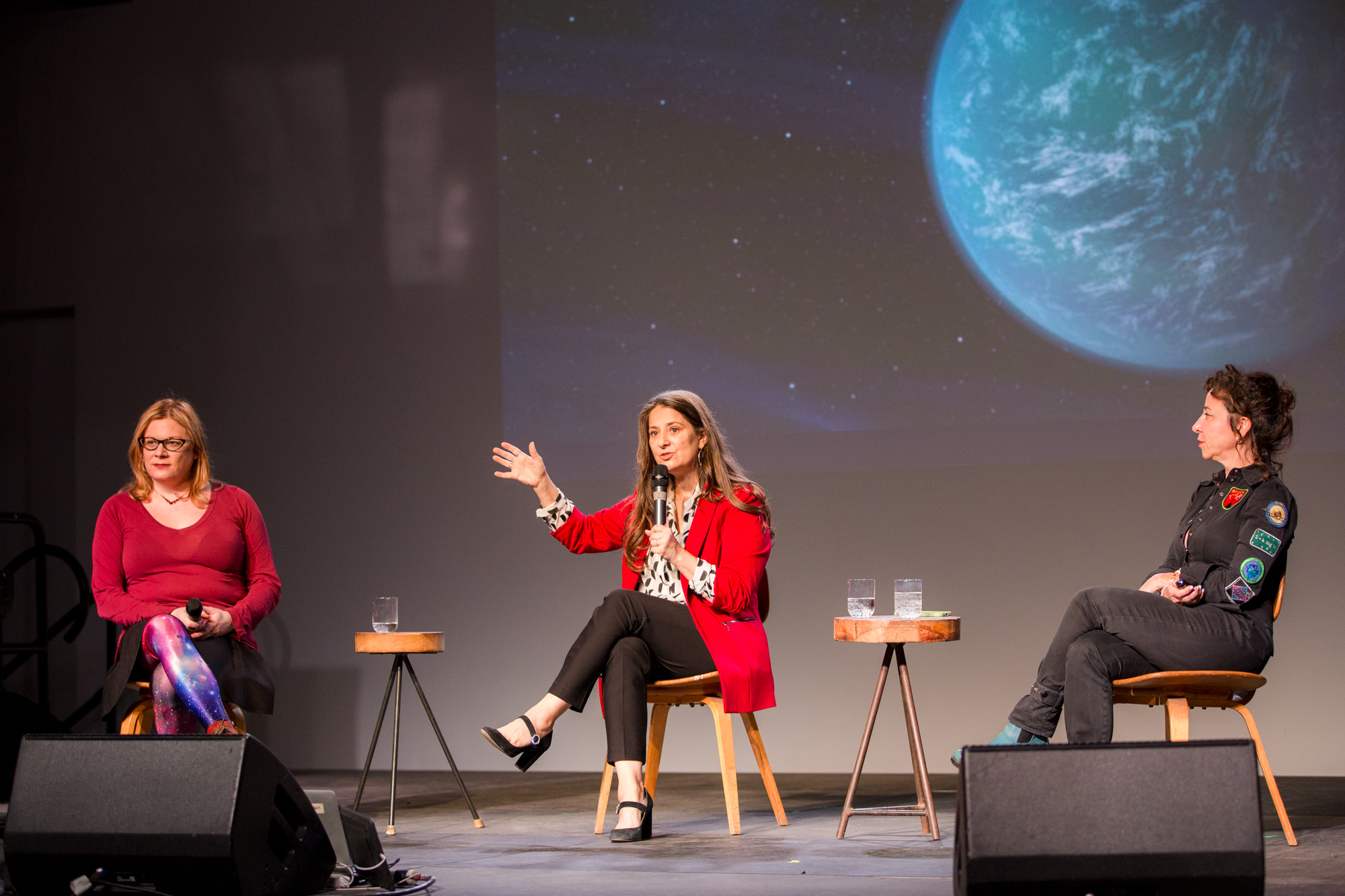Three scientists sit against the backdrop of a blue planet at a panel discussion. The scientist in the center, Natalie Batalha, speaks into a microphone.
