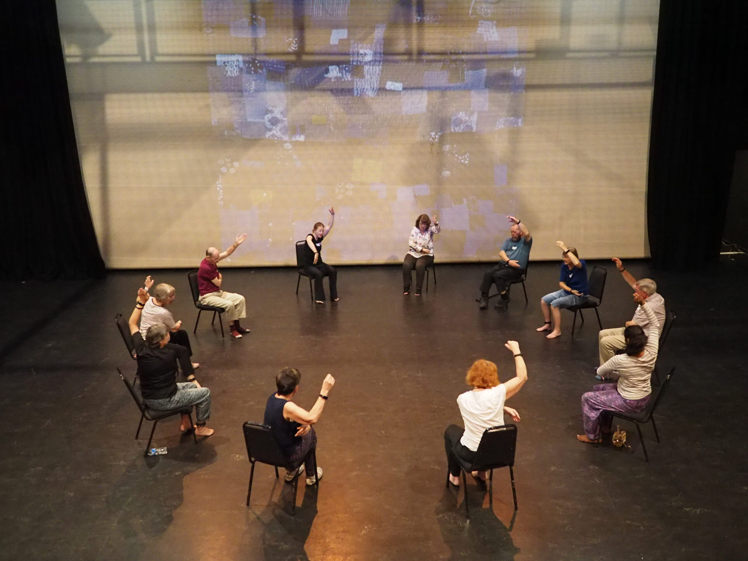 A group of people with Parkinson's disease sit together in a circle with one arm raised, dancing.