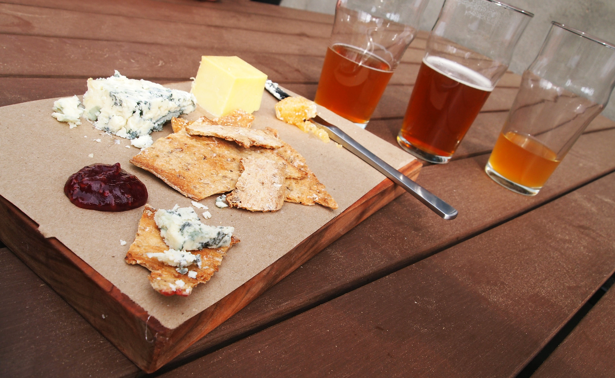 Three types of cheese, including blue cheese, sit on a charcuterie board with crackers. Three different half-full glasses of beer sit on the table in the background.