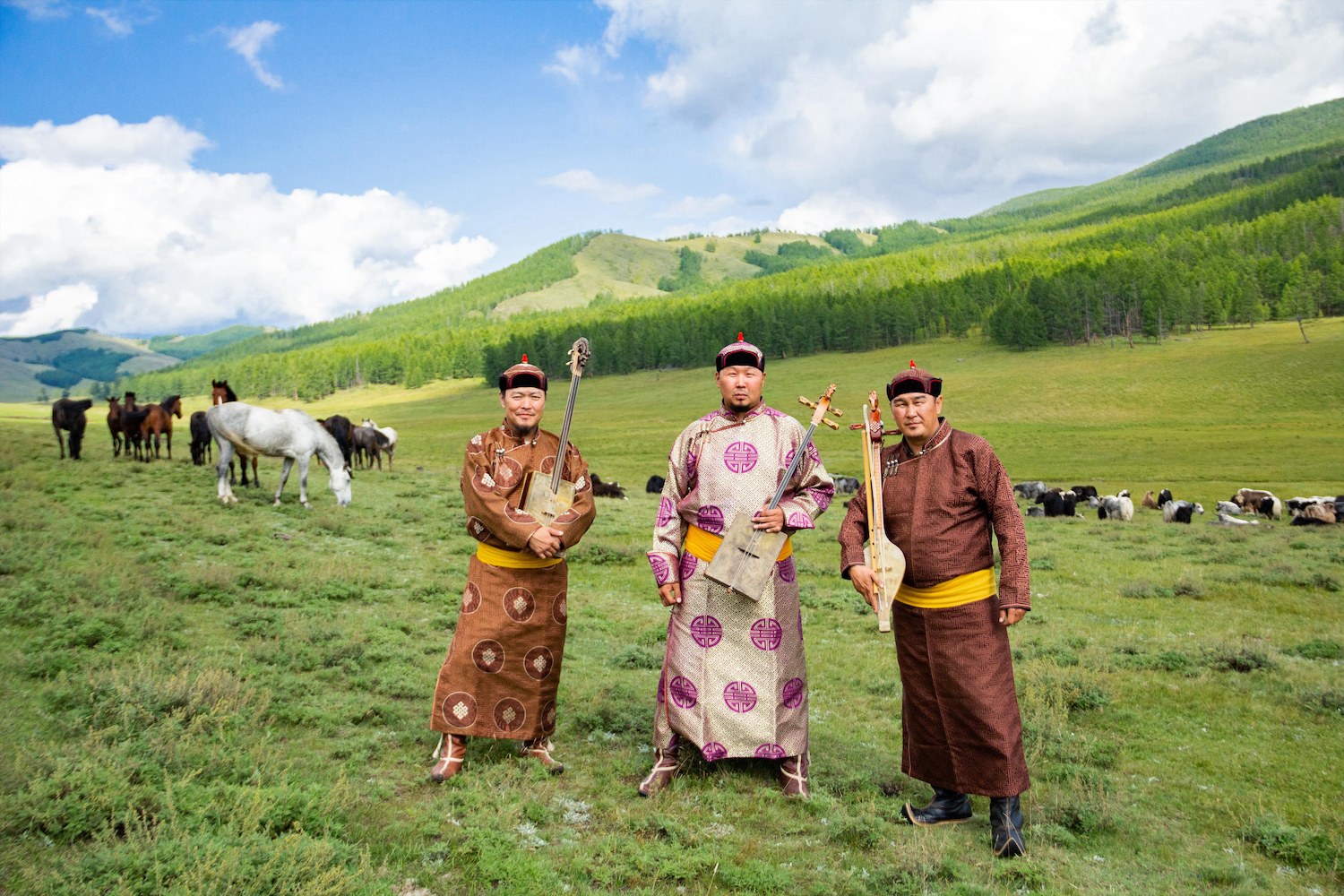 Three Tuvan men, the members of Alash, stand in a green field holding instruments. Horses graze in the background.