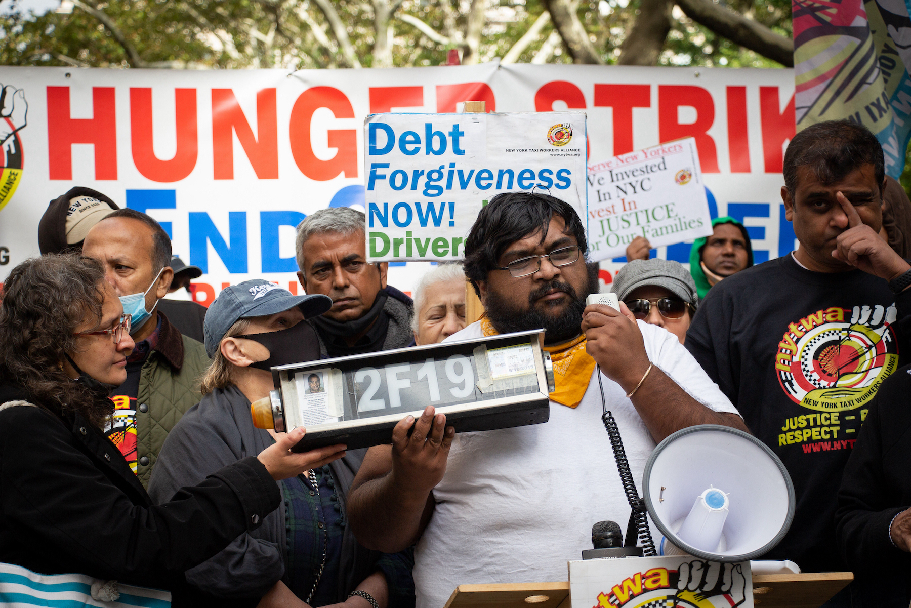 A crowd of people are protesting in a park, with banners reading "Hunger strike" and "Debt forgiveness now!" A young man at the front of the crowd speaks into a megaphone while holding a taxi medallion number.