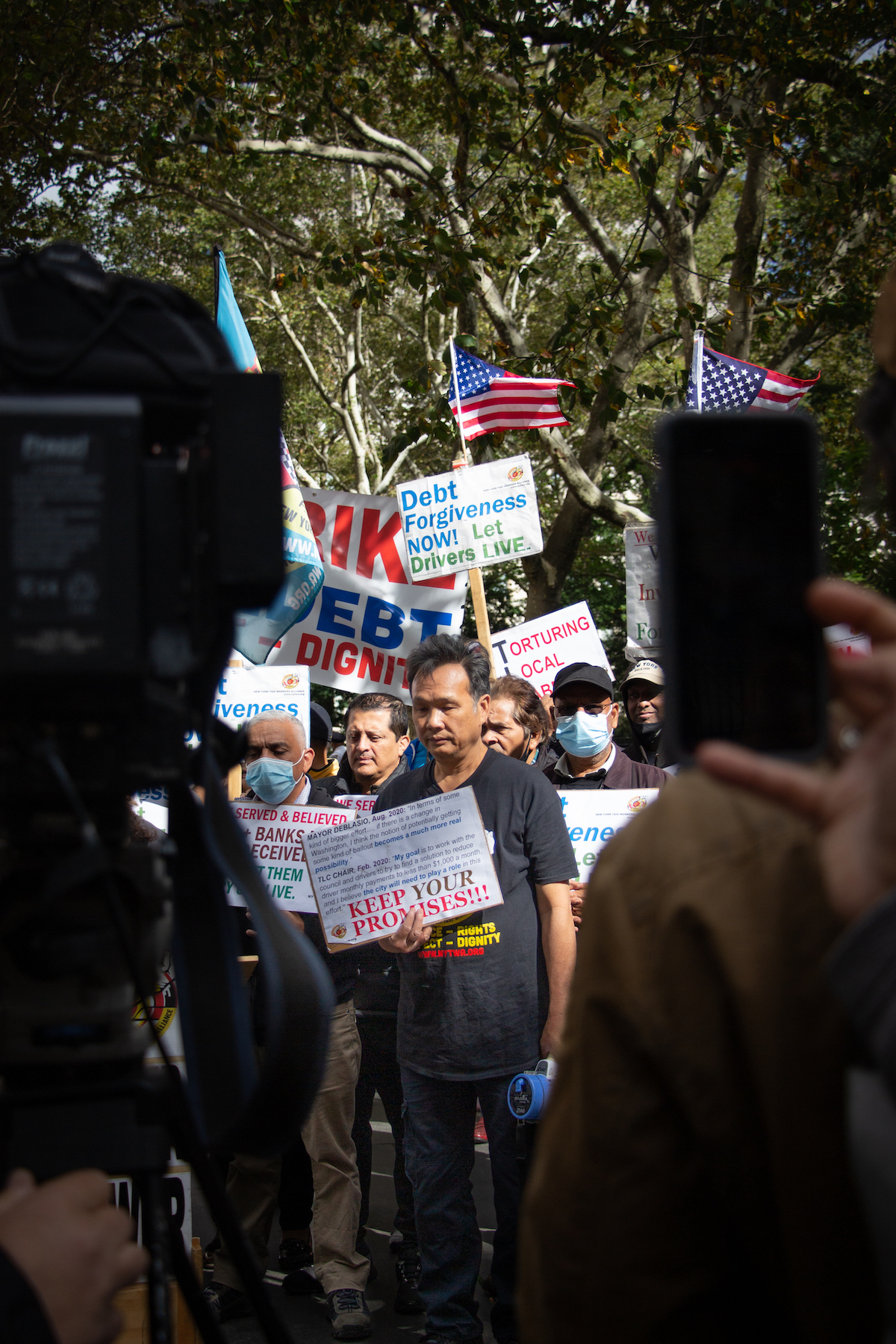 A crowd of protestors holding signs stand in a park. One older man in the center of the group, his eyes closed, is framed by cameras in the foreground.