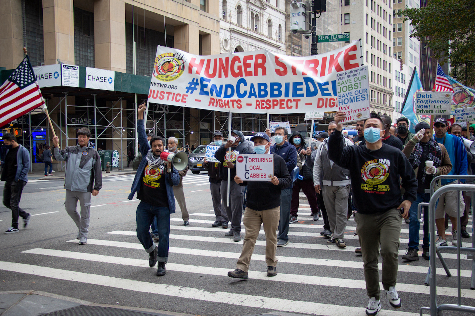 A group of people march on a city street holding protest signs. The biggest one reads "Hunger Strike: #EndCabbieDebt." Many of the marchers wear matching sweatshirts with the logo of the New York Taxi Workers Alliance.