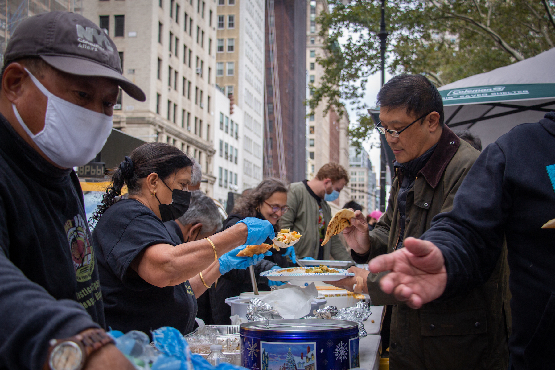 People on one side of a long table set outside on a New York City street dish out food on paper plates to people on the other side.