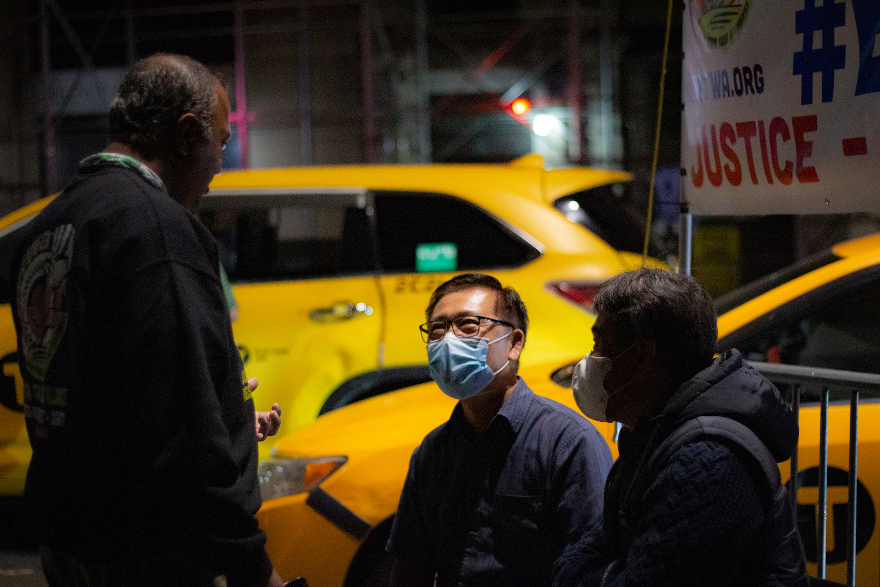 Three older men are talking on a dark New York City sidewalk, with yellow cabs parked in the street behind them. Two of the men are sitting on plastic folding chairs, and one has his face (with a medical face mask) illuminated by the street lights.