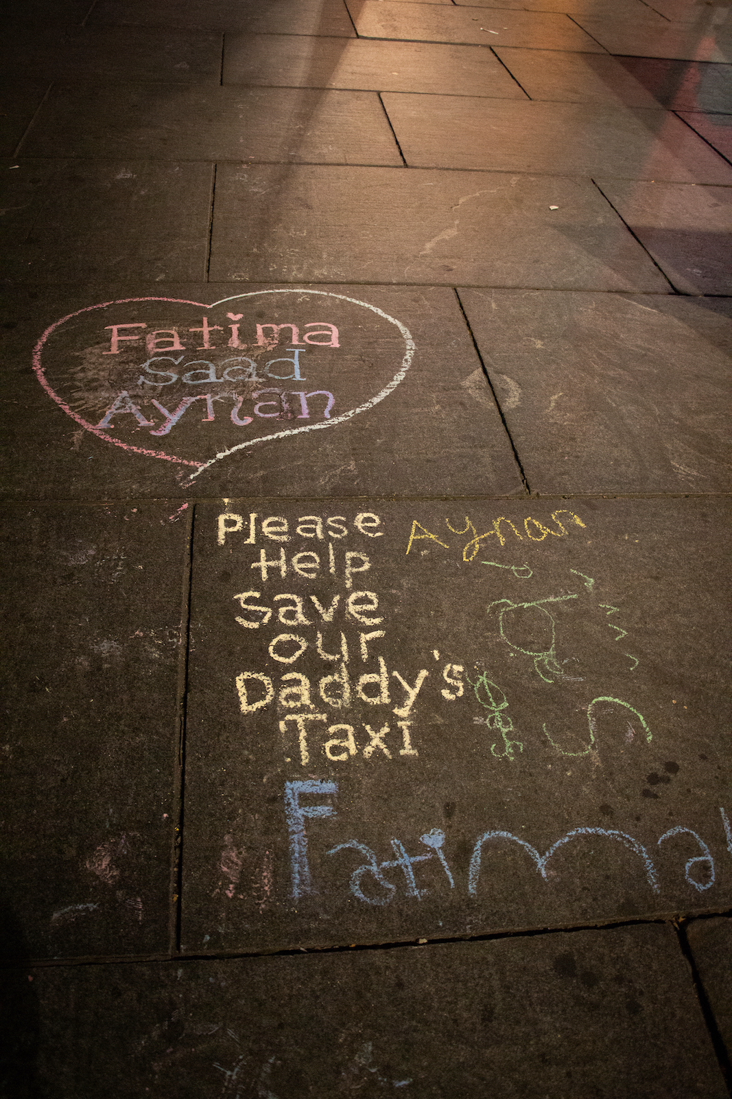 The colorful chalk drawings and writings of children are lit by streetlights on a dark city sidewalk. Inside a heart are the names Fatima, Saad and Aynan, and a message underneath the heart reads "Please help save our Daddy's taxi."
