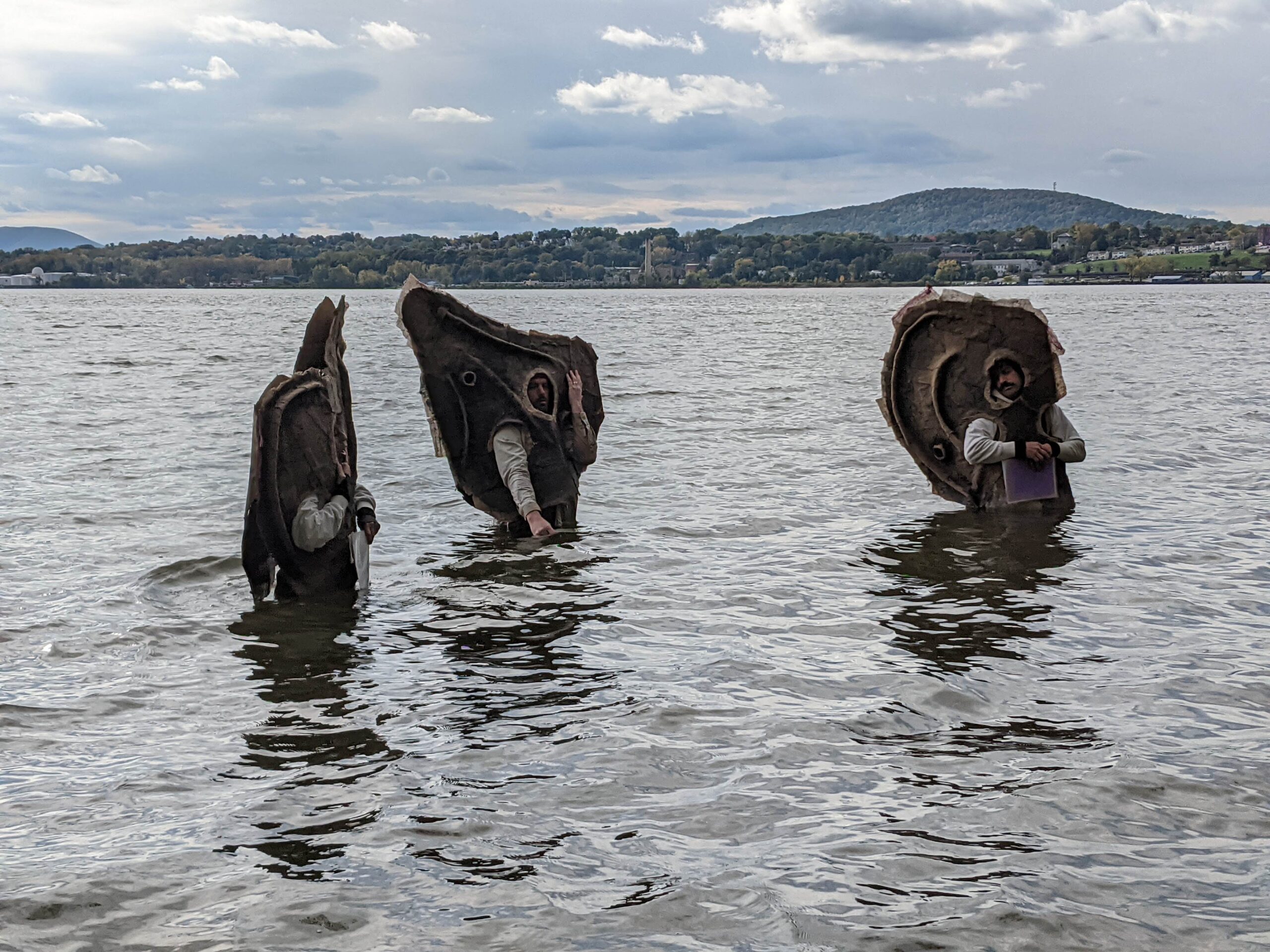 Three actors waist deep in the Hudson River wearing cardboard cutouts shaped like oyster shells.