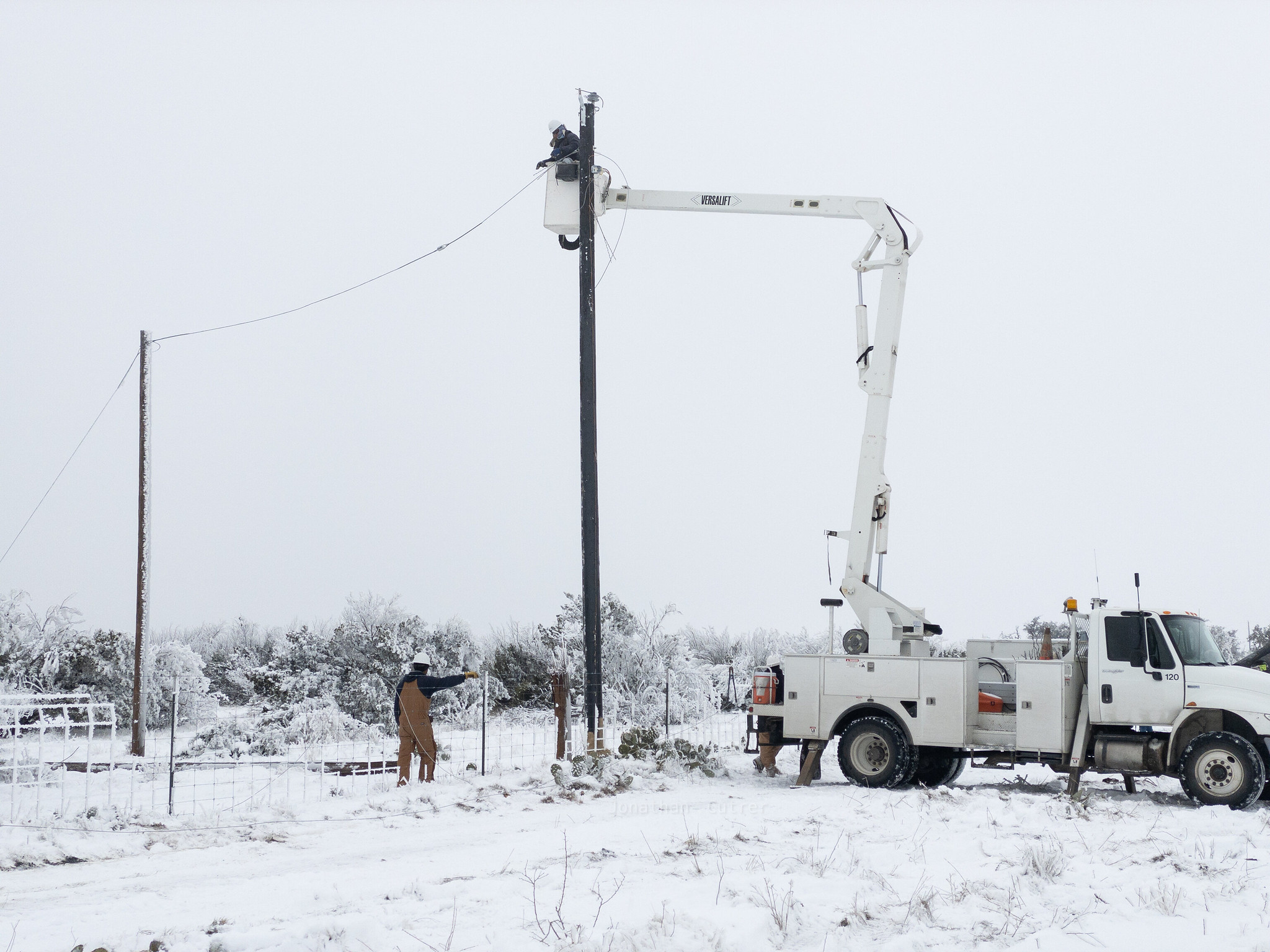 A lineman is working at the top of an electric wire pole while a colleague stands below in the snow.