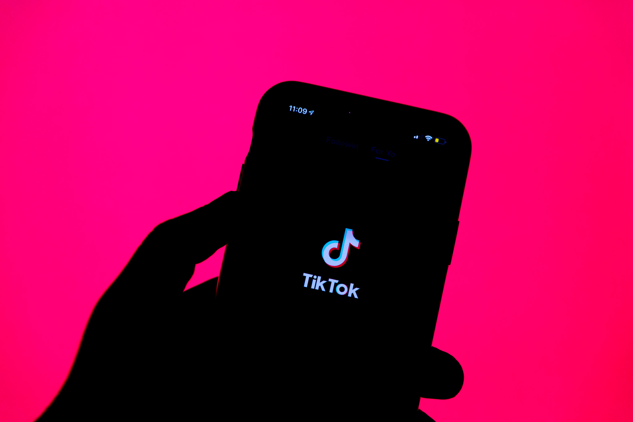 The silhouette of a hand holds an iPhone up against a bright, pink background. The phone displays the TikTok logo.