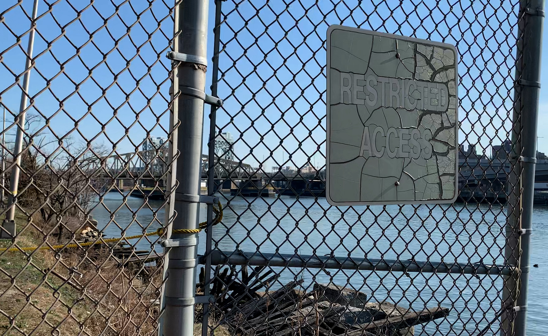 The waterfront along the Harlem River behind a chain link fence. A sign reads "Restricted Access"