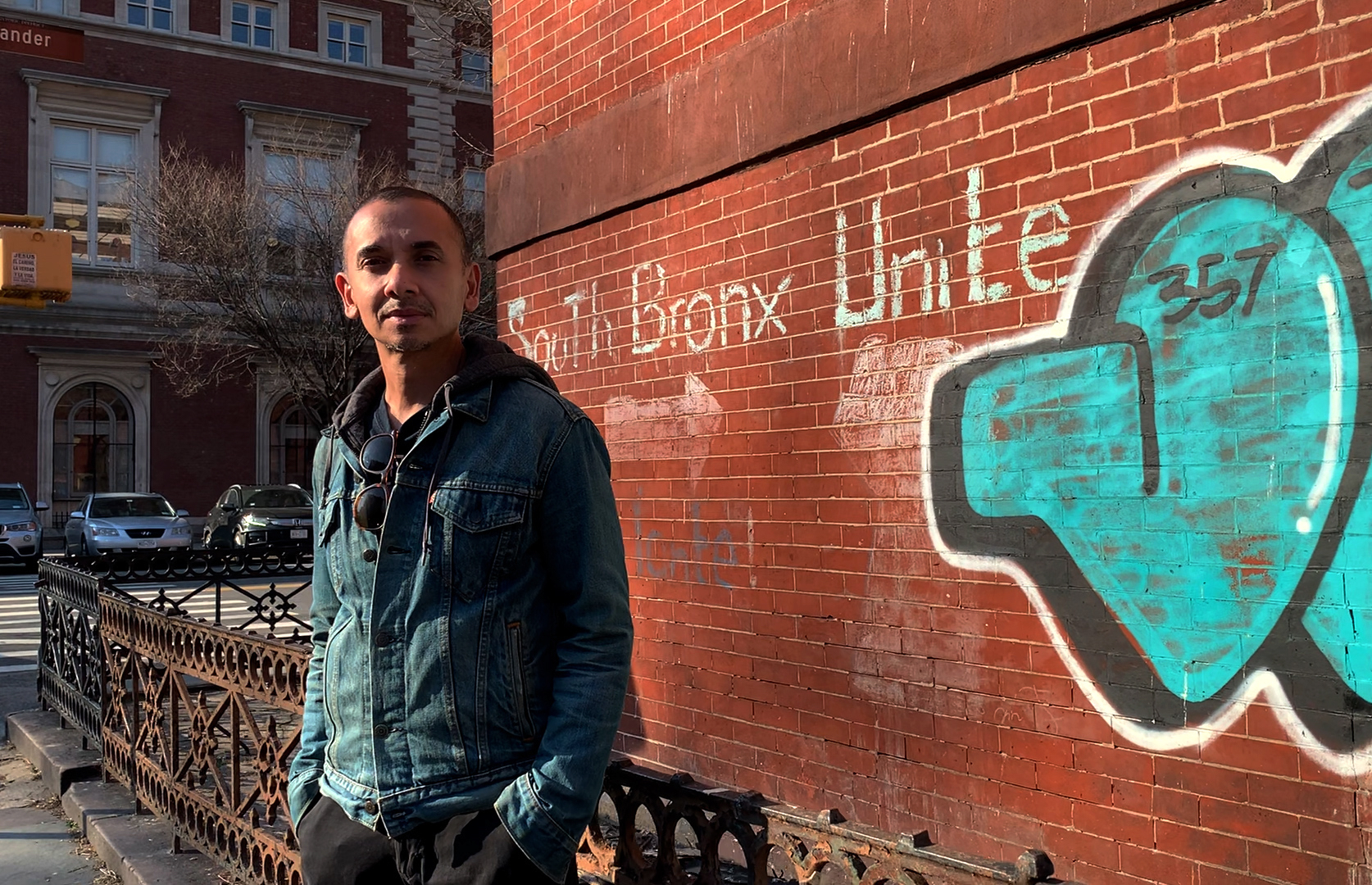 Arif Ullah stands on a street corner in front of a brick wall with South Bronx Unite written in chalk on the wall