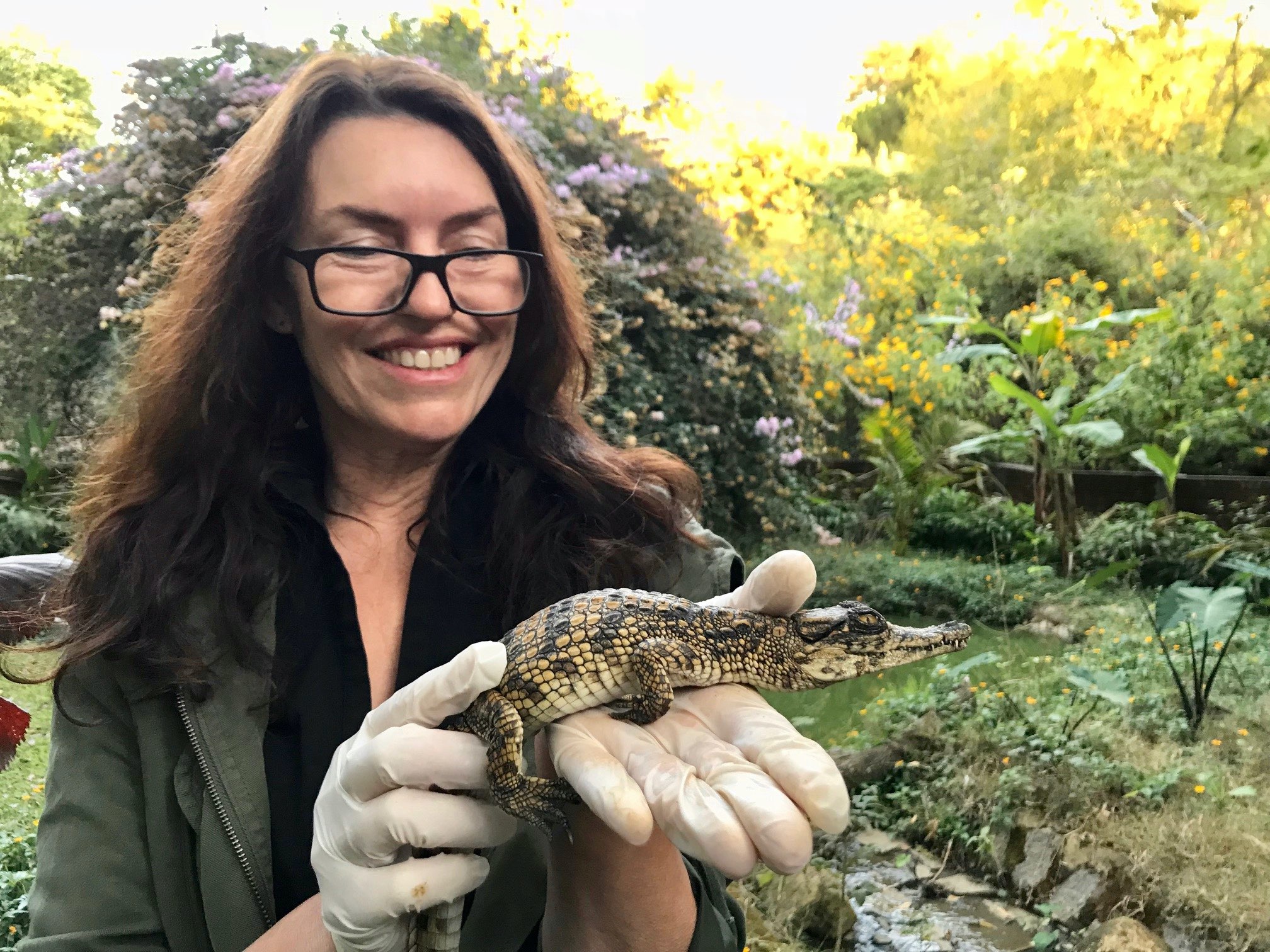 Evon Hekkala holds a baby crocodile horizontally in gloved hands. She's smiling and wearing black glasses. In the background there are lush trees and flowers.