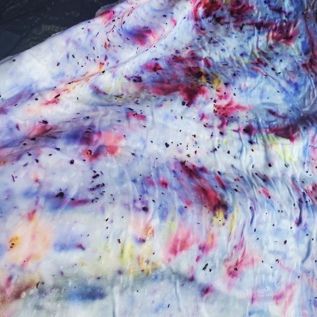 A wrinkled white sheet of fabric is dappled and smattered with varying shades of purple, red, and yellow. Some of the shades bleeds into one another like watercolors, while others are opaque pigmented flecks.