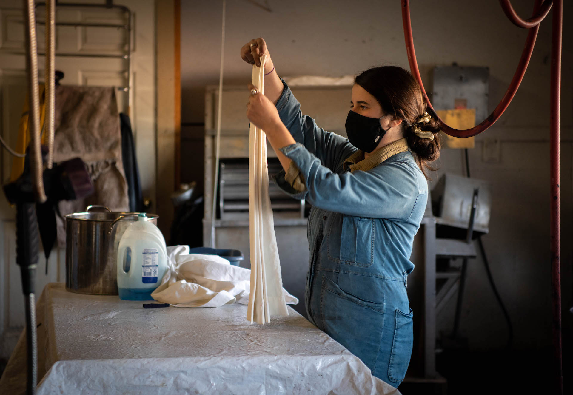 Winona Quigley stands in the frame in her denim coveralls, folding and holding taut a piece of cream colored fabric over a plastic covered workbench. A large stainless steel pot and a large tub of blue detergent sit on the far end of the work bench.
