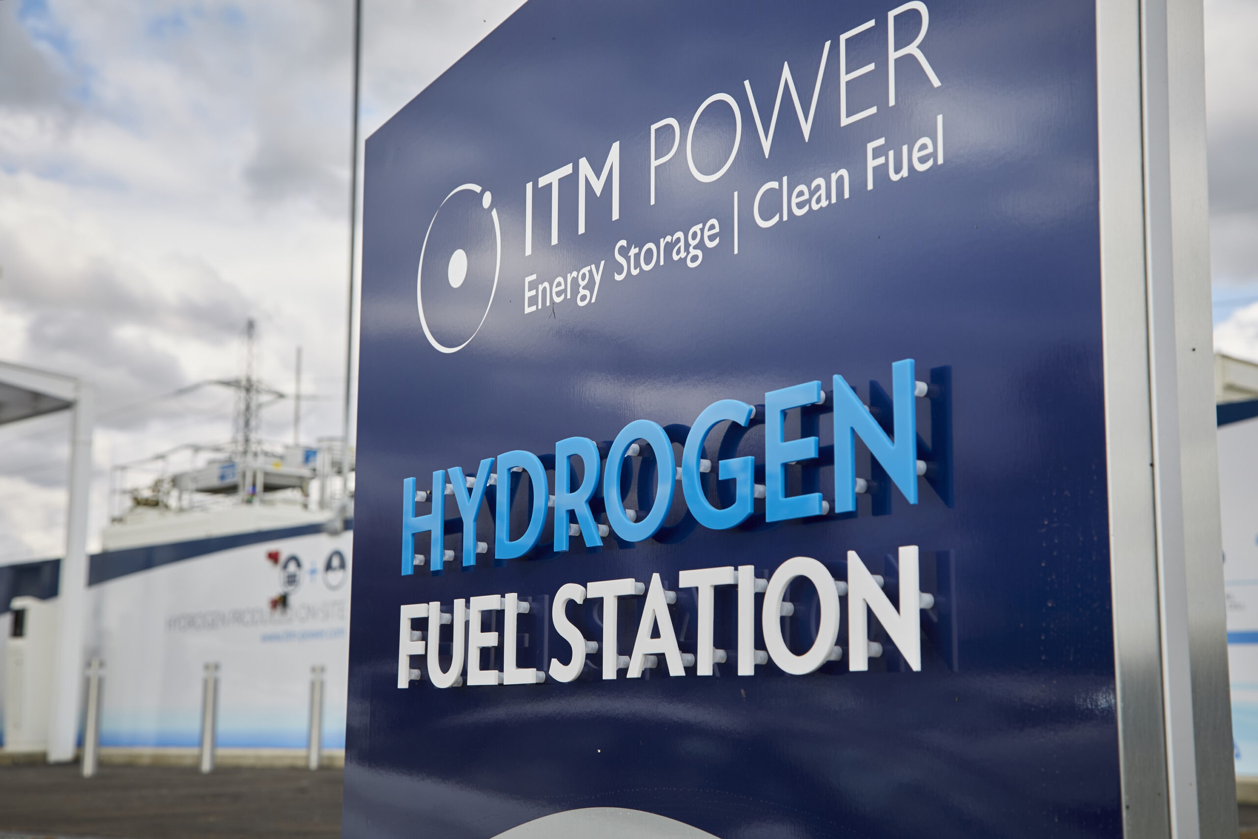 A picture of a blue sign with white lettering outside of a power plant. The sign says "ITM Power. Energy Storage | Clean Fuel. Hydrogen Fuel Station."