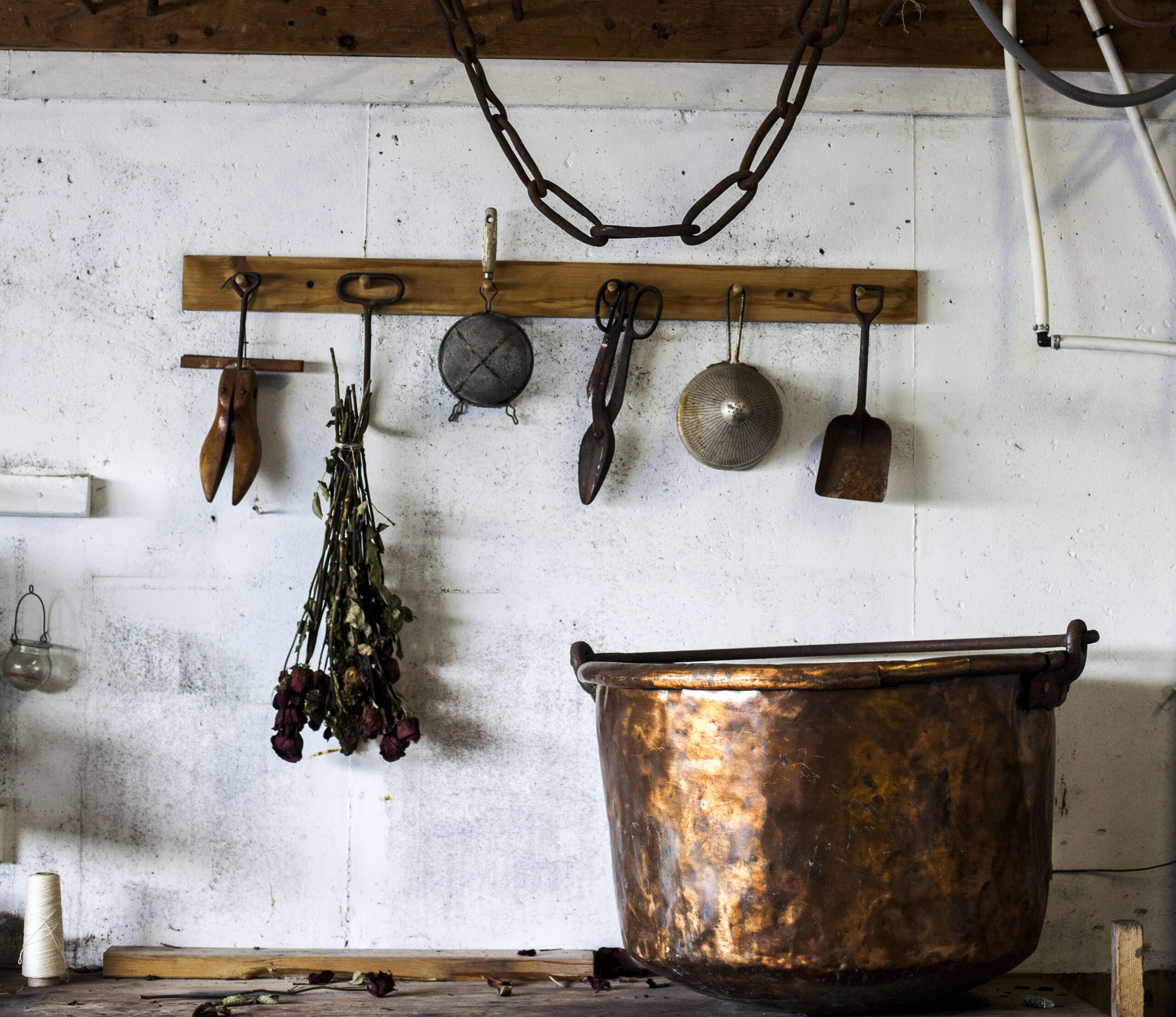 A large dappled copper bowl sits atop a wooden countertop. Above it sits a rack with hooks for different tools and materials, including sieves of three different sizes, two pairs of sheers, and a bouquet of red dried flowers. 