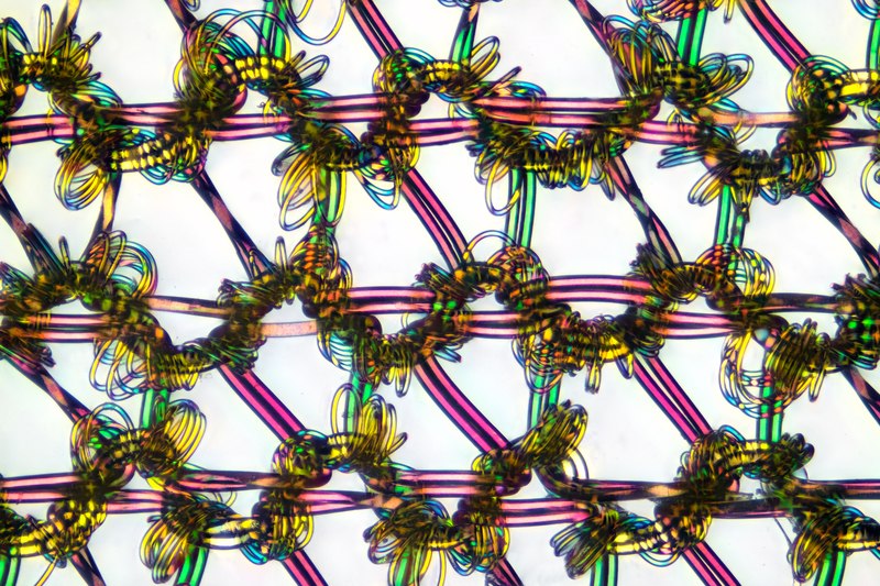 A microscope image of a nylon stocking, showing a network of trapezoidal pink plastic fibers connected and reinforced by coils of green and yellow fibers.