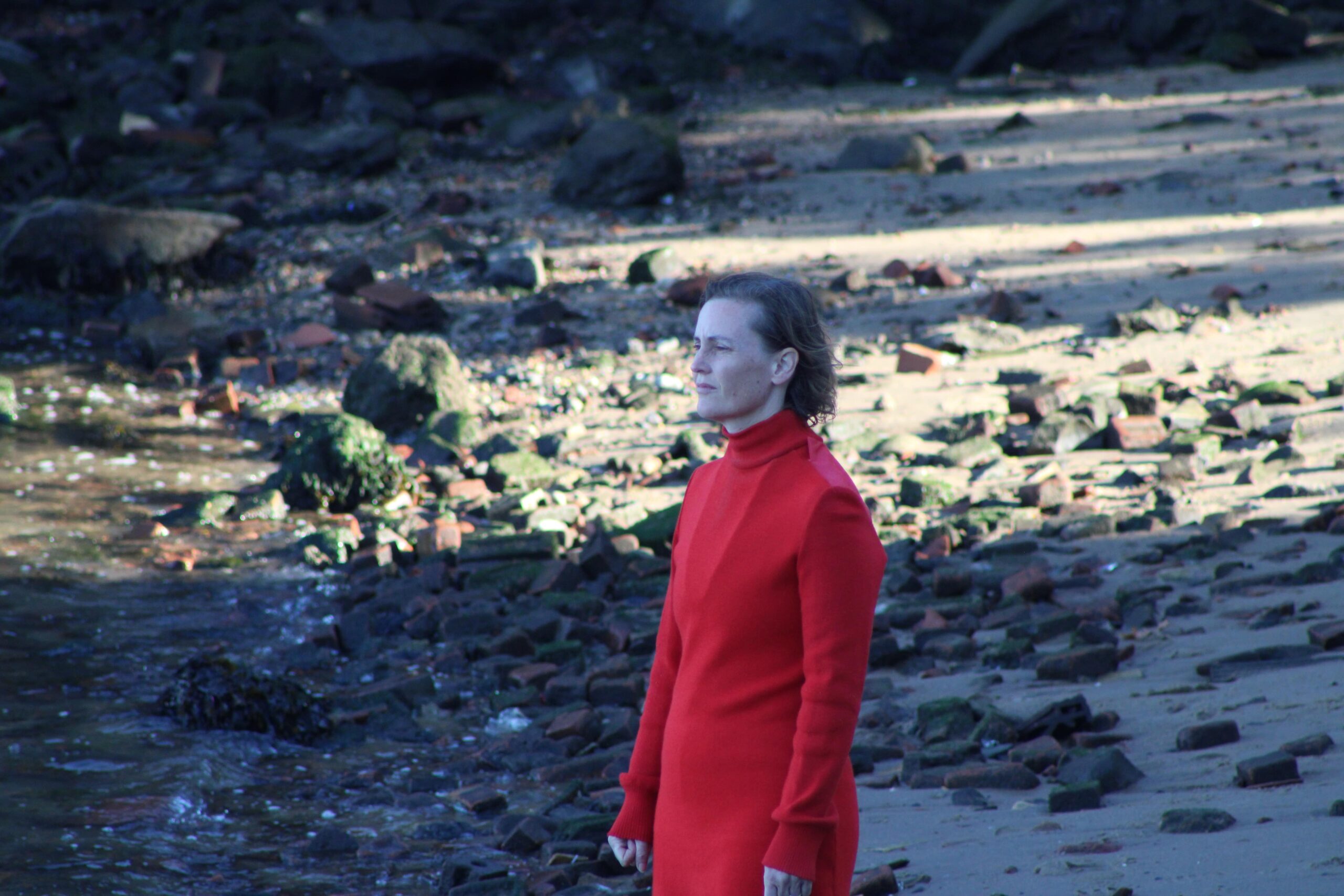 A photograph of the activist/artist Sarah Cameron Sunde in a red wind breaker, standing on the beach in her climate change performance.