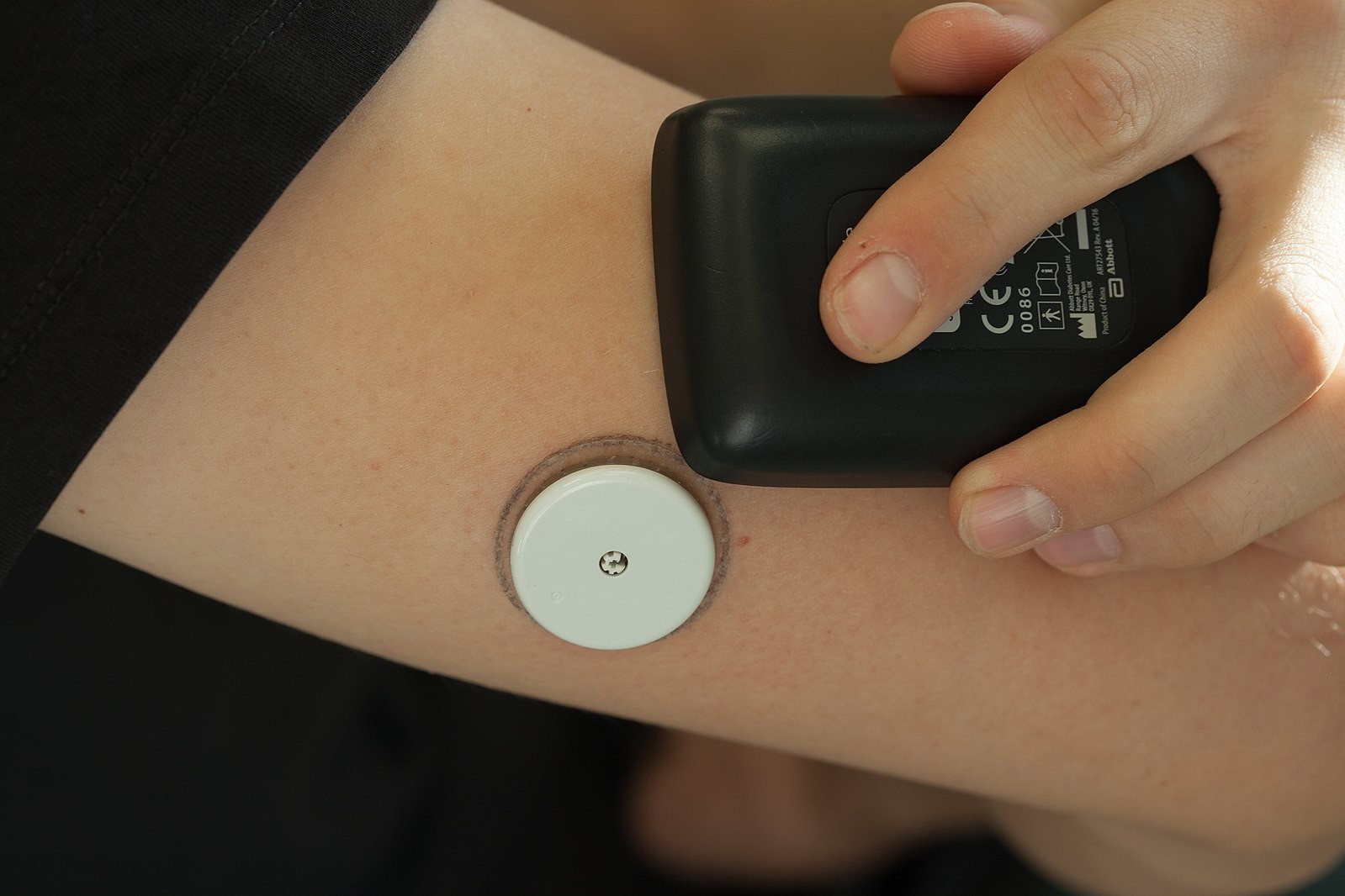 Person with a white, circular continuous glucose monitor on the back of their upper arm holding black display monitor next to it