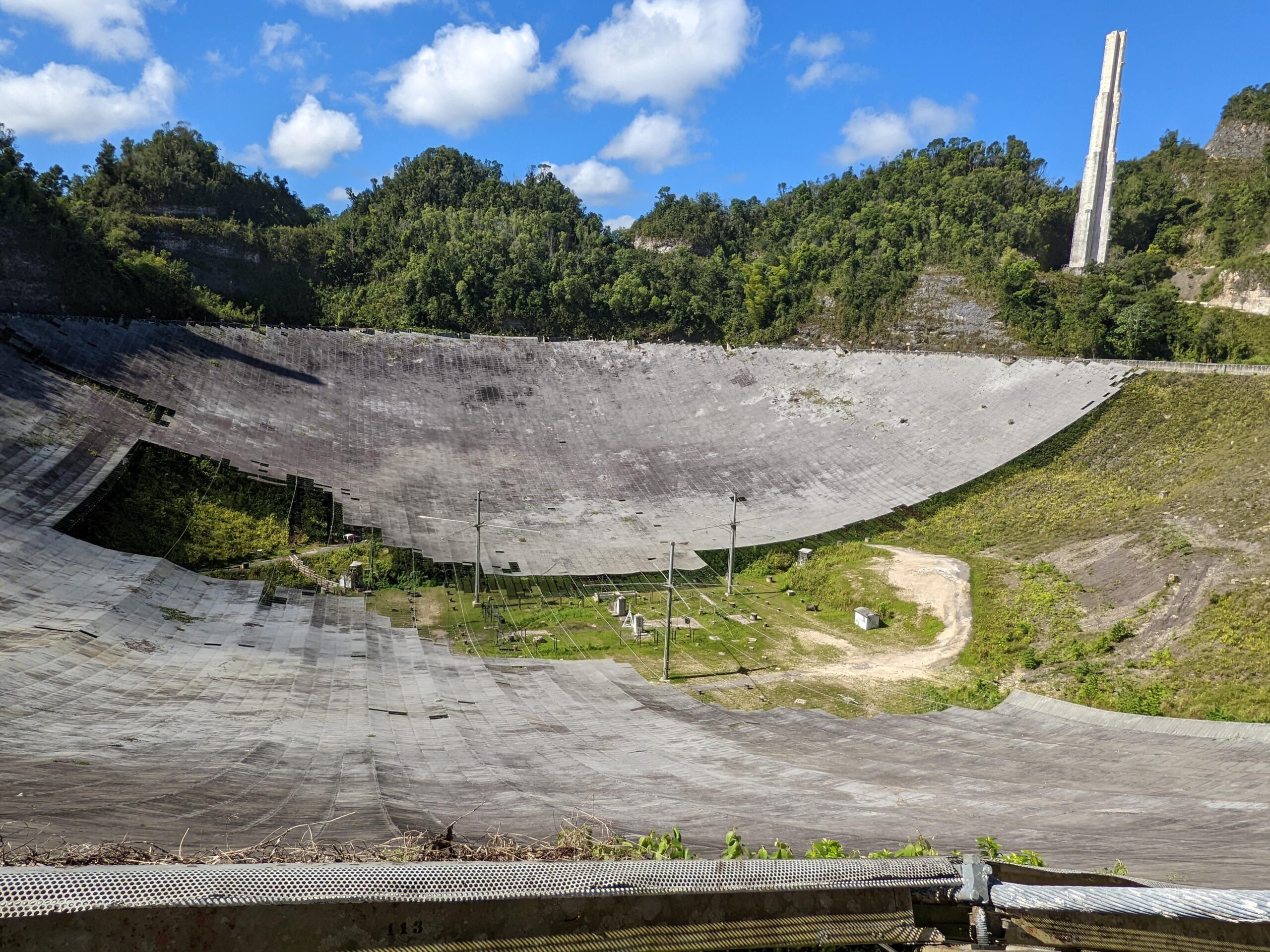 The remains of the Arecibo Telescope dish as viewed from the rim. About 40% of the grey dish is missing, revealing the green foliage underneath