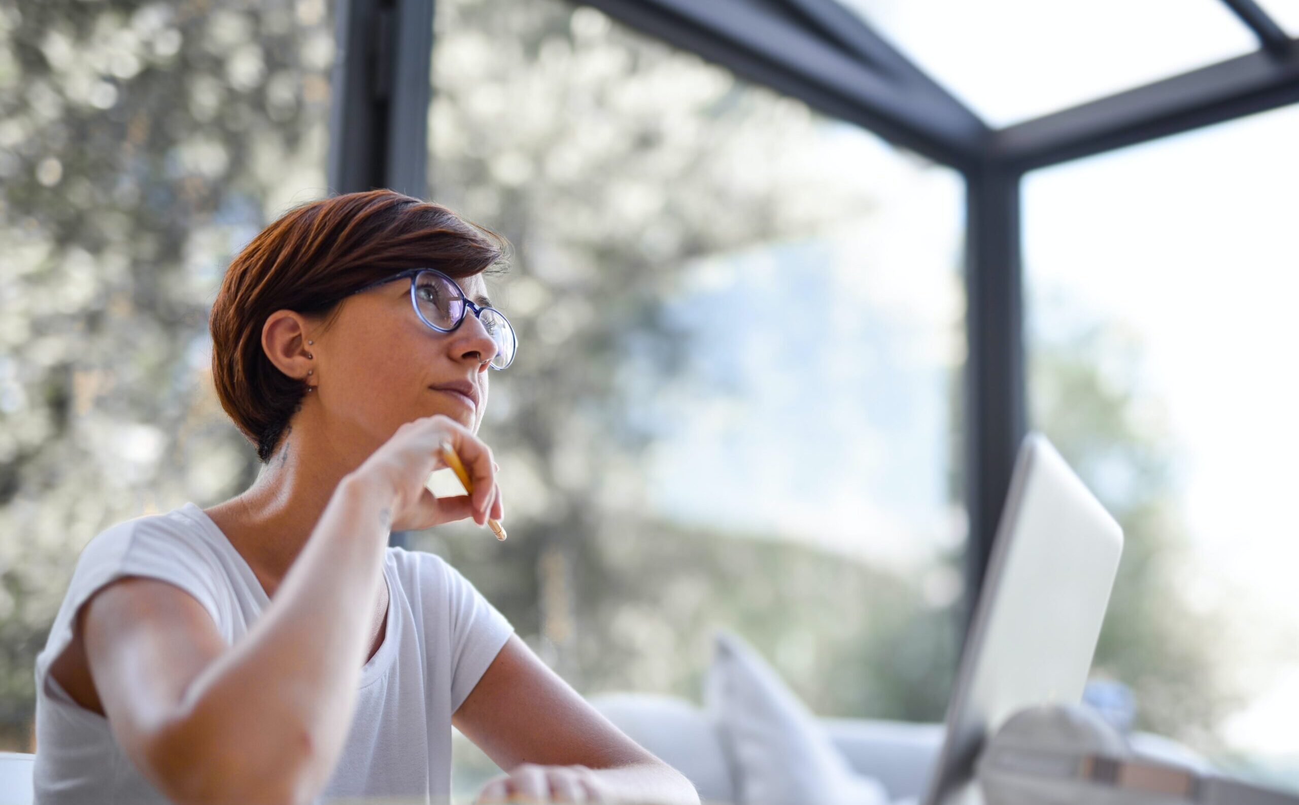 person with short hair and glasses working at a computer surrounded by windows, thinking pensively