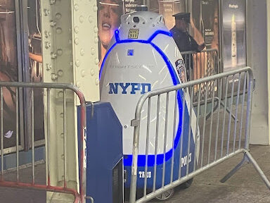 A blue and white robot with NYPD written on it, behind an indoor barrier in the subway