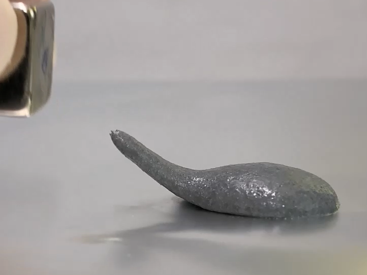 A grey metallic putty is on a table, a strand of it reaches out towards a silvery cube in the top left corner