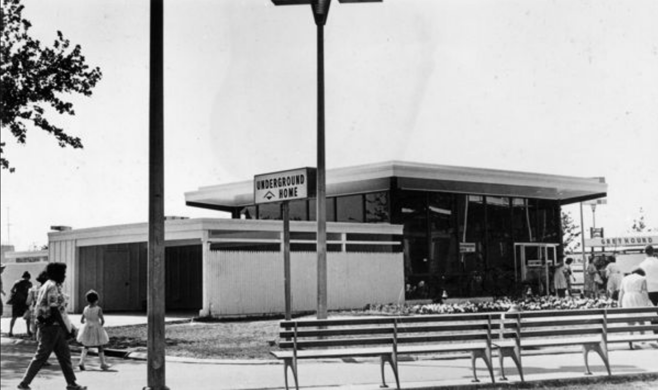 A black and white photo of a park with two benches facing square buildings with a sign "The Underground Home"