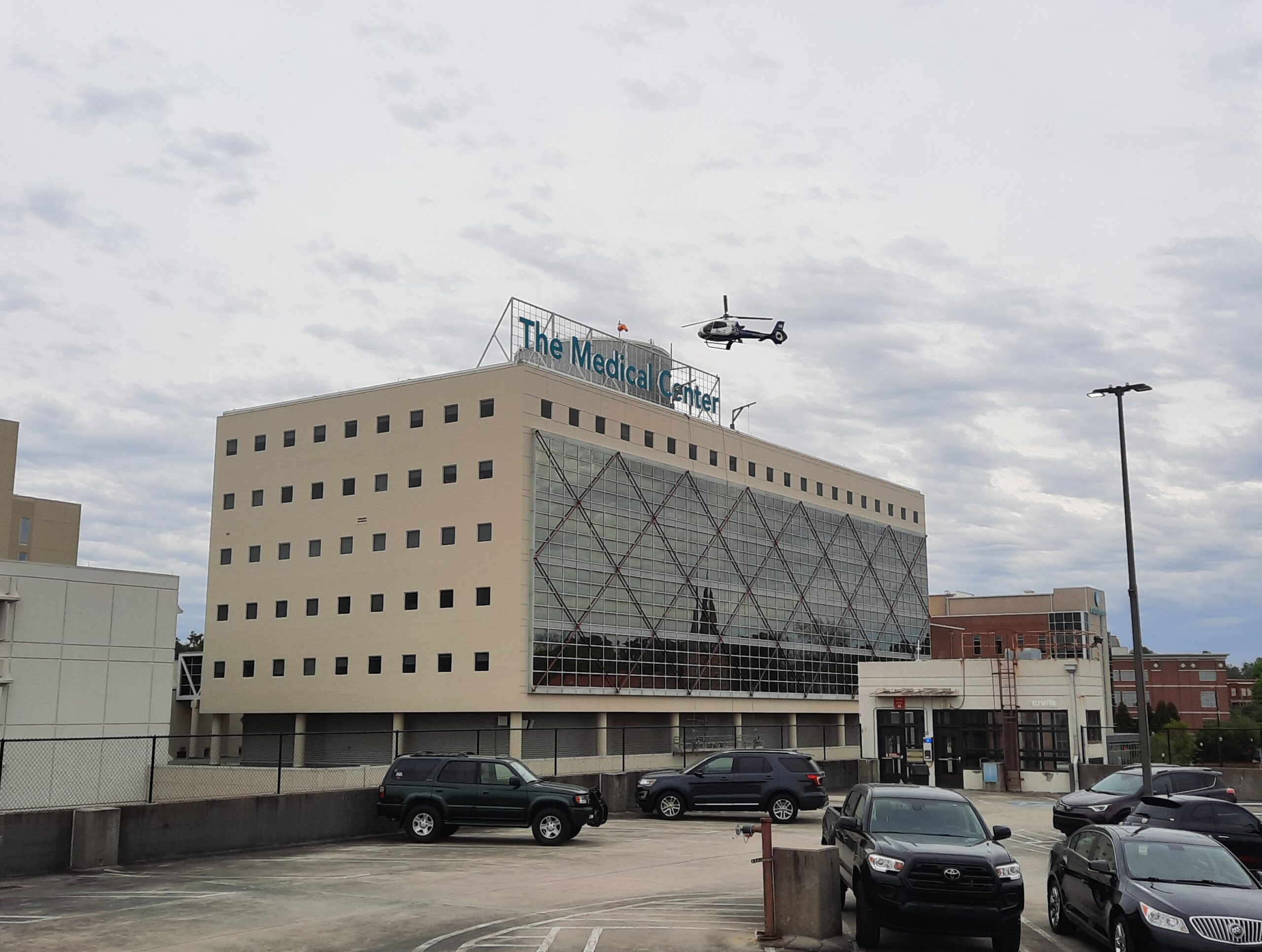 A hospital building with cars parked out front and a helicopter flying overhead.