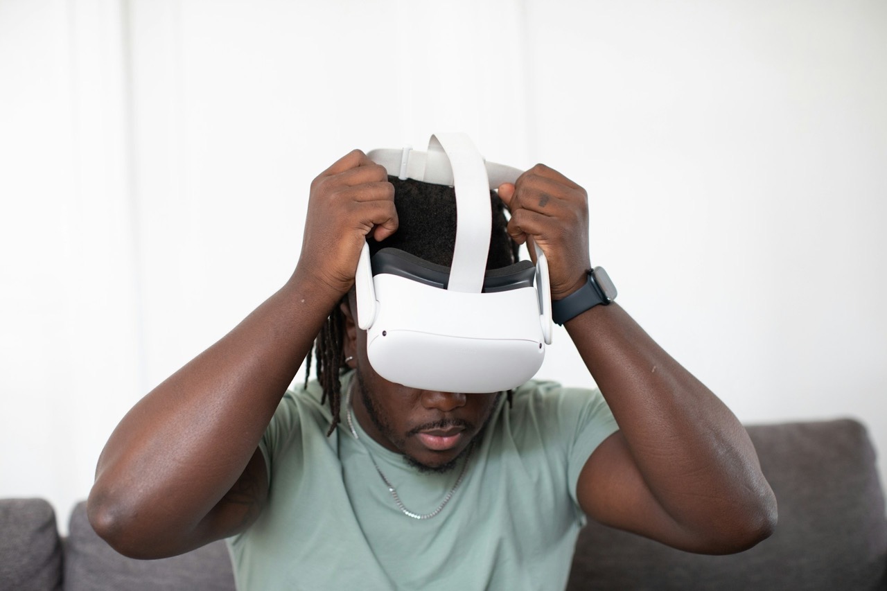 A man sits on a gray couch and puts on a white VR headset, like the type some are using to treat schizophrenia