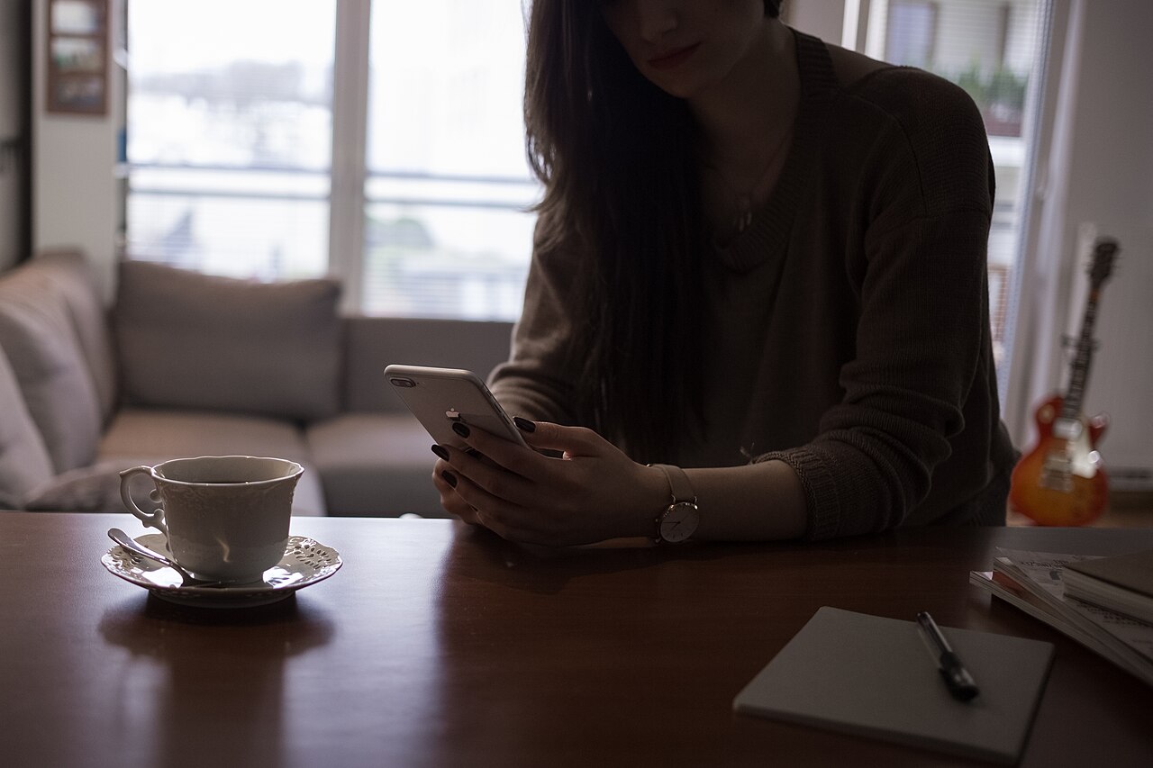 An anonymous woman sits at her a desk indoors, holding an iPhone, with a small cup and saucer to her right.