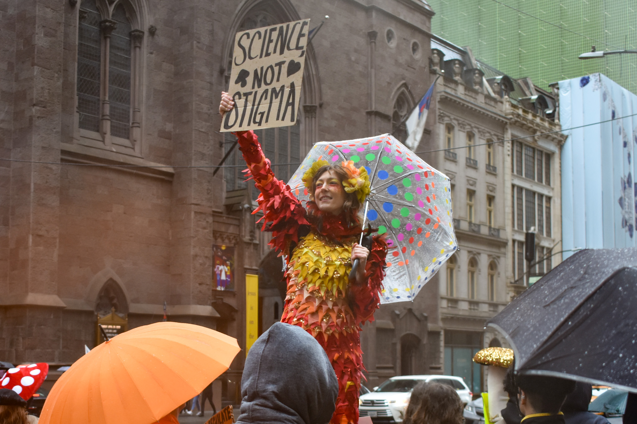 A woman wearing psychedelic colors and holding an umbrella stands on stilts with a sign that reads SCIENCE NOT STIGMA